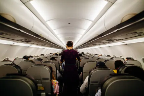 A flight attendant in a plum uniform walks down the aisle of a full airplane cabin, seen from behind