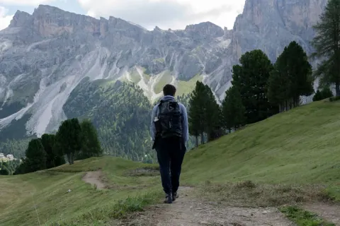 A hiker with a black backpack walks a dirt trail through a green alpine meadow toward towering limestone peaks