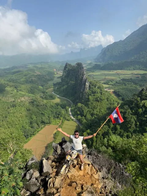 Merald on a rocky limestone viewpoint above Vang Vieng, Laos, arms wide and holding a Lao flag over a valley of green karsts and rice fields